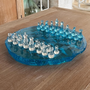 Könnte beinhalten: Ein rundes Schachspiel mit einem durchscheinenden blauen Harzbrett mit gezacktem, unebenem Rand und einem Gittermuster. Das Set enthält zwei verschiedene Gruppen von klaren und blau getönten Glasschachfiguren, darunter Könige, Damen, Läufer, Springer, Türme und Bauern, die auf der Spielfläche angeordnet sind.