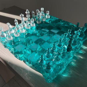Può includere: Un set di scacchi in vetro trasparente con una scacchiera turchese. I pezzi degli scacchi trasparenti sono disposti su una scacchiera a scacchi. La scacchiera ha una superficie strutturata e irregolare, che le conferisce un aspetto unico. Il set è esposto su una superficie chiara.