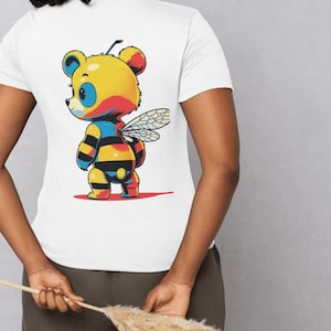 Puede incluir: Camiseta blanca con un diseño de oso-abeja de dibujos animados. El oso-abeja tiene una cabeza amarilla, ojos azules y un cuerpo a rayas negro y amarillo. Tiene alas y está sobre una superficie roja. La camiseta es de manga corta.