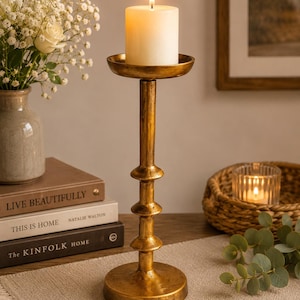 May include: A tall, gold-toned metal candle holder with a textured, weathered finish. The stand features a circular base, a slender central column with three decorative horizontal rings, and a wide, flat top plate holding a single white pillar candle.