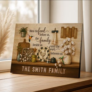 May include: Wooden family sign with a rustic design. The sign features the text "Bless the food before us, the family beside us, and the love between us." Decorative elements include a rooster plate, hummingbird, and cotton stems.