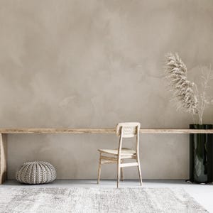 May include: A minimalist interior with a long wooden table, a woven chair, and a grey ottoman. A tall black vase holds dried plants, and a patterned rug covers the floor. The wall is a solid, muted beige colour.