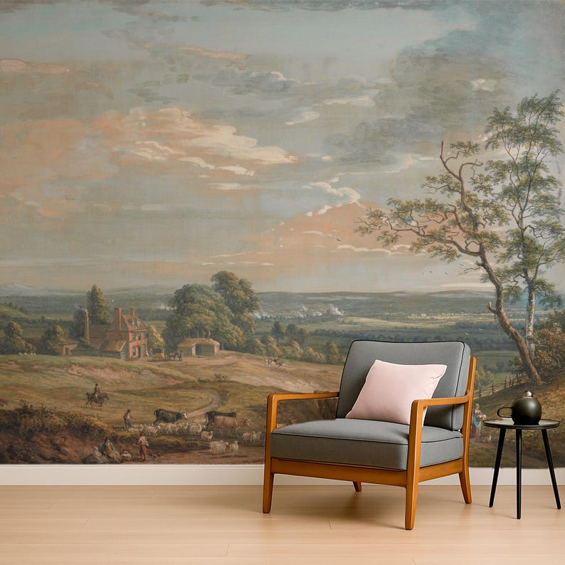 Puede incluir: Un sill&oacute;n gris tapizado con brazos de madera y un coj&iacute;n rosa claro se sit&uacute;a frente a un gran mural de pared que representa un paisaje pastoral con colinas, una casa de ladrillo, ganado pastando y un gran &aacute;rbol bajo un cielo nublado al atardecer.
