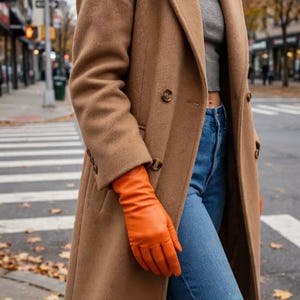 May include: A camel-colored wool coat with a double-breasted button front, paired with a gray cropped top and blue denim jeans. The outfit is accented by bright orange leather gloves that extend past the wrist, creating a bold color contrast.