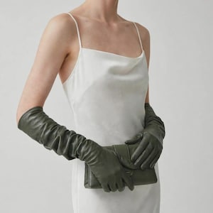 Peut inclure: Une robe nuisette blanche à fines bretelles associée à de longs gants en cuir vert olive qui arrivent aux coudes. Les gants présentent une texture froncée et plissée le long des avant-bras et sont portés en tenant une pochette assortie.