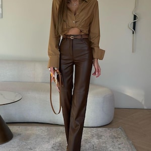 May include: A woman's outfit featuring a brown button-down shirt with a knotted front, a brown leather belt, and brown leather pants. The outfit is completed with brown pointed-toe boots and a brown purse with a patterned scarf.