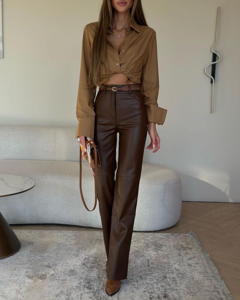 May include: A woman's outfit featuring a brown button-down shirt with a knotted front, a brown leather belt, and brown leather pants. The outfit is completed with brown pointed-toe boots and a brown purse with a patterned scarf.