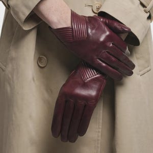Può includere: Un paio di guanti in pelle color bordeaux intenso con finitura liscia e un distinto dettaglio a coste a forma di V nella zona del polso. I guanti sono indossati con un trench beige con bottoni visibili e colletto.
