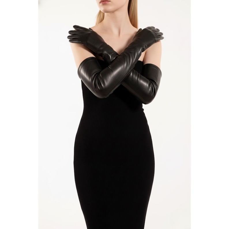 May include: Black leather opera gloves, extending past the elbows, are crossed over the chest. The gloves are smooth and form-fitting. The model is wearing a black dress. The background is plain white.