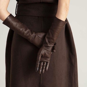 Peut inclure: Une paire de longs gants en cuir marron foncé qui dépassent le poignet, présentant une construction lisse et ajustée avec des détails de couture subtils le long des doigts et des paumes, conçus pour couvrir entièrement les mains et les avant-bras.