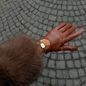 Könnte beinhalten: Ein brauner Lederhandschuh mit Handgelenksaussparung, getragen mit einer goldfarbenen analogen Armbanduhr mit weißem Zifferblatt und dünnem Metallarmband, kombiniert mit einem dunkelbraunen Kunstfellärmel, der über das Handgelenk reicht.