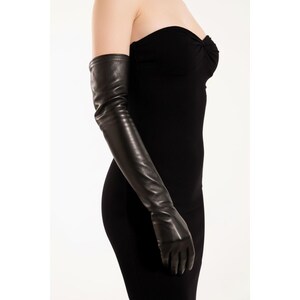 May include: A pair of long, black leather gloves extending past the elbow. The gloves are sleek and form-fitting, with a smooth texture. The image shows the gloves worn with a black strapless dress, creating a sophisticated look.