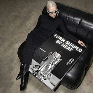 May include: A black and white poster with the text "FORM SHAPED BY HEAT" and a chrome-like sculpture. The poster is held by a person wearing a black dress and sunglasses. The poster's design features a grid background and the words "CHROME STUDY".