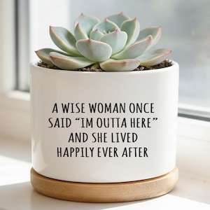 Op de afbeelding: Witte keramische plantenpot met een vetplant. De pot heeft de tekst "A WISE WOMAN ONCE SAID 'IM OUTTA HERE' AND SHE LIVED HAPPILY EVER AFTER". Hij staat op een ronde houten basis.