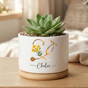 Op de afbeelding: Een witte keramische plantenbak met een vetplant. De plantenbak heeft een hartvormig stethoscoopontwerp met gele bloemen en de naam "Chloe" in script. De plantenbak staat op een ronde houten basis.