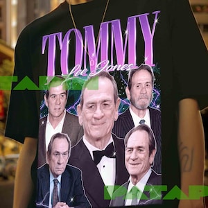 May include: Black t-shirt featuring a graphic of Tommy Lee Jones with the text "TOMMY Lee Jones" in a purple and white design. The graphic includes multiple images of the actor. The shirt is worn by a person.