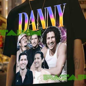 May include: Black t-shirt with the name "DANNY" in a rainbow gradient font. The shirt features a collage of images of people. The background is a blurred city scene.