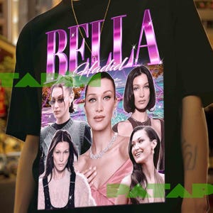 Peut inclure: T-shirt noir avec le nom "BELLA" dans une police rétro rose et violette, et "Hadid" en dessous. Le t-shirt présente plusieurs images de Bella Hadid. L'arrière-plan montre une scène de ville.