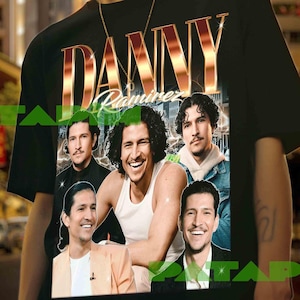 May include: Black t-shirt featuring a graphic of Danny Ramirez with the name in gold lettering. The shirt showcases multiple images of the actor. The model is wearing a gold chain and bracelet. The background is a city street.
