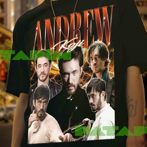 Pode incluir: T-shirt preto com um gráfico de vários homens e o nome "ANDREW" em uma fonte retrô. A camiseta é usada por uma pessoa com um colar de corrente dourada. O fundo é uma paisagem urbana.