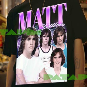 Puede incluir: Camiseta negra con un gráfico de Matt Dillon, el nombre "MATT" en una fuente retro púrpura. El gráfico incluye múltiples imágenes del actor. La camiseta la lleva una persona frente a un fondo de ciudad borroso.
