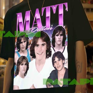 Puede incluir: Camiseta negra con un gráfico de Matt Dillon, el nombre "Matt Dillon" en una fuente retro púrpura y múltiples imágenes de Matt Dillon. La camiseta la lleva una persona.