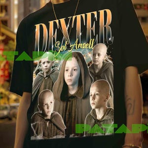 Könnte beinhalten: Schwarzes T-Shirt mit dem Wort "DEXTER" in einem Farbverlauf und "Sol Ansell" darunter. Das Shirt zeigt eine Collage von Kindern in Kapuzenmänteln. Der Hintergrund ist eine Stadtstraße.