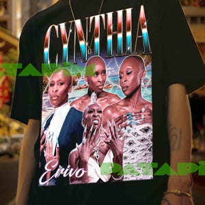 Puede incluir: Camiseta negra con un gráfico de Cynthia Erivo y el nombre "CYNTHIA" en una fuente retro colorida. La imagen incluye múltiples retratos de Erivo. La camiseta la lleva una persona frente a un fondo urbano borroso.