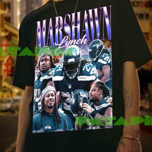 Puede incluir: Camiseta negra con un gráfico de Marshawn Lynch con uniforme de fútbol. El diseño incluye el nombre "Marshawn Lynch" en fuente morada y blanca. La camiseta muestra múltiples imágenes del jugador de fútbol.