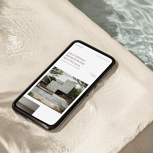 May include: A smartphone with a black frame displays a website with the address "928 LUNEDALE MISSION ROAD SEATTLE 98123". The website features a modern architectural design. The phone rests on a light-colored, textured surface near water.