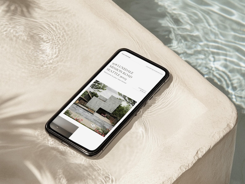 May include: A smartphone with a black frame displays a website with the address "928 LUNEDALE MISSION ROAD SEATTLE 98123". The website features a modern architectural design. The phone rests on a light-colored, textured surface near water.