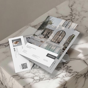 Op de afbeelding: Een stapel vastgoedmarketingmaterialen, waaronder brochures en visitekaartjes, met afbeeldingen van een modern huis. De brochures tonen architectonische details en de prijs van het pand. De visitekaart bevat een foto en contactgegevens.