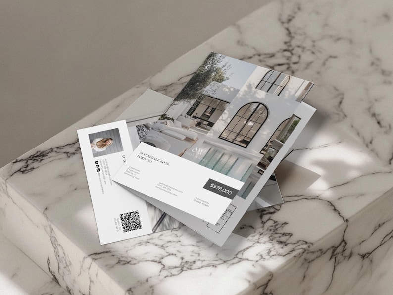Op de afbeelding: Een stapel vastgoedmarketingmaterialen, waaronder brochures en visitekaartjes, met afbeeldingen van een modern huis. De brochures tonen architectonische details en de prijs van het pand. De visitekaart bevat een foto en contactgegevens.