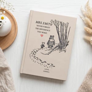 May include: A beige hardback book featuring a black line drawing of Winnie the Pooh and Piglet walking near trees. The text reads MRS. SMITH, NEVER FORGET THE DIFFERENCE YOU MAKE, and LOVE, SOPHIA, with a small pink heart icon centred on the cover.