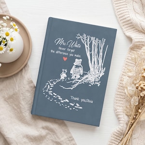 Puede incluir: Un libro de tapa dura color azul pizarra con un dibujo de líneas blancas de dos osos caminando hacia unos árboles. El texto dice: Mrs. White, Never forget the difference you make, y Thank you, Olivia. Un pequeño corazón rojo está entre las líneas.