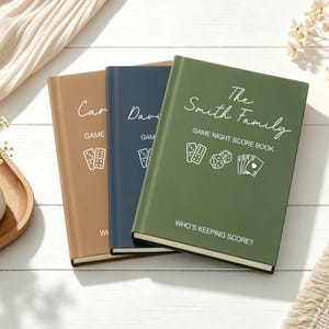 May include: Three hardback game night score books in tan, navy blue, and olive green. Each book features white text including the title Game Night Score Book, icons of dominoes, dice, and playing cards, and the phrase Who's Keeping Score?