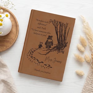 Op de afbeelding: Een lichtbruin notitieboek met een leren kaft en een zwarte lijntekening van Winnie de Poeh en Knorretje op een pad. De kaft bevat het citaat "It makes such a difference," said Pooh, "To have someone who believes in you," met de naam "Mrs. Jones" en "Thank you, Sophia" eronder gedrukt.