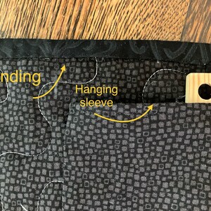 May include: Close-up of a quilted textile corner. The fabric features a dark gray square pattern with white stitching. A black patterned binding frames the edge. A hanging sleeve is visible, along with a wooden hanger.