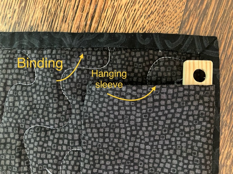May include: Close-up of a quilted textile corner. The fabric features a dark gray square pattern with white stitching. A black patterned binding frames the edge. A hanging sleeve is visible, along with a wooden hanger.
