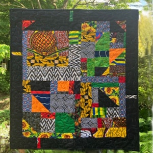 May include: A colorful patchwork quilt with a black border. The quilt features various patterned fabrics, including sunflowers, geometric shapes, and a pineapple design. The quilt is hanging outdoors, with green foliage in the background.