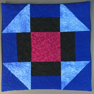 May include: A quilted pillow with a geometric design. The pillow is made of blue, black, and red fabric. The center of the pillow is a large red square surrounded by four black squares. The corners of the pillow are made of blue fabric with a white pattern.