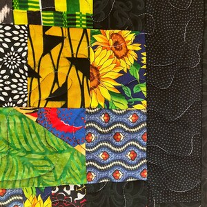 May include: Close-up of a patchwork quilt with a variety of fabric patterns. The quilt features squares of black, yellow, green, and blue fabrics with floral, geometric, and abstract designs. White stitching outlines the squares.