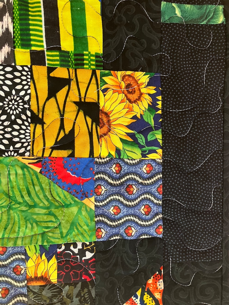 May include: Close-up of a patchwork quilt with a variety of fabric patterns. The quilt features squares of black, yellow, green, and blue fabrics with floral, geometric, and abstract designs. White stitching outlines the squares.