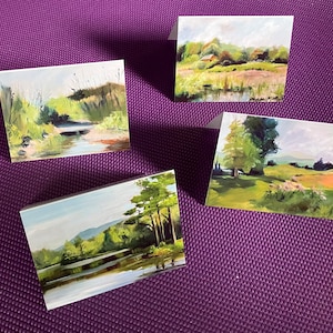 May include: Four landscape art greeting cards. Each card features a different painting of a natural scene, including a river, a field, and a lake. The paintings use a palette of greens, blues, and browns, with visible brushstrokes. The cards are displayed on a purple textured surface.