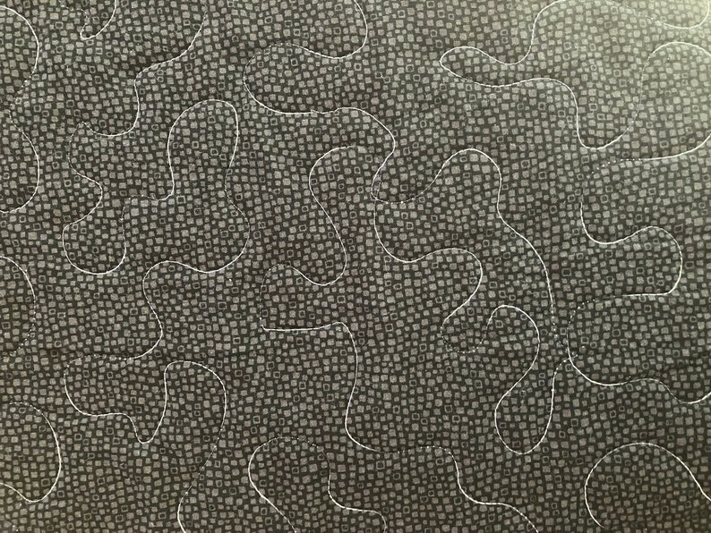 May include: Close-up of a quilted fabric with a dark gray background featuring a pattern of small squares. White thread creates a flowing, organic design across the surface. The quilting adds texture and visual interest to the textile.