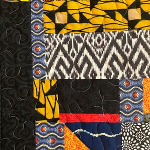 May include: Close-up of a patchwork quilt with various fabric patterns. The quilt features sections of black, yellow, orange, blue, and white fabrics. The fabrics have floral, geometric, and abstract designs. White stitching outlines the patterns.