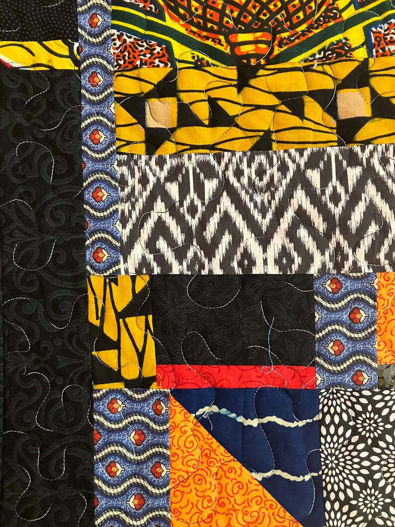 May include: Close-up of a patchwork quilt with various fabric patterns. The quilt features sections of black, yellow, orange, blue, and white fabrics. The fabrics have floral, geometric, and abstract designs. White stitching outlines the patterns.