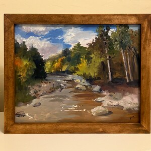 May include: Oil painting of a river scene with a wooden frame. The artwork depicts a flowing river surrounded by trees and rocks, under a blue sky with white clouds. The painting is framed in a natural wood.