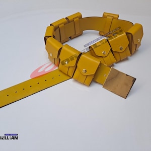 Op de afbeelding: Mosterdgele utility riem met meerdere zakjes en een rechthoekige gesp. De riem heeft een reeks kleine, vierkante zakjes met goudkleurige knoopsluitingen. De riem is gemaakt van een glad, leerachtig materiaal.