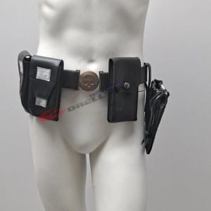 Op de afbeelding: Zwarte gereedschapsriem met meerdere zakjes en een gesp in de vorm van een schedel. De riem heeft twee vierkante zakjes met zilveren accenten, een centraal zakje en een holster. De riem wordt op een mannequin getoond.